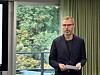 Mann i mørk jakke står innendørs med papirer foran et stort vindu.